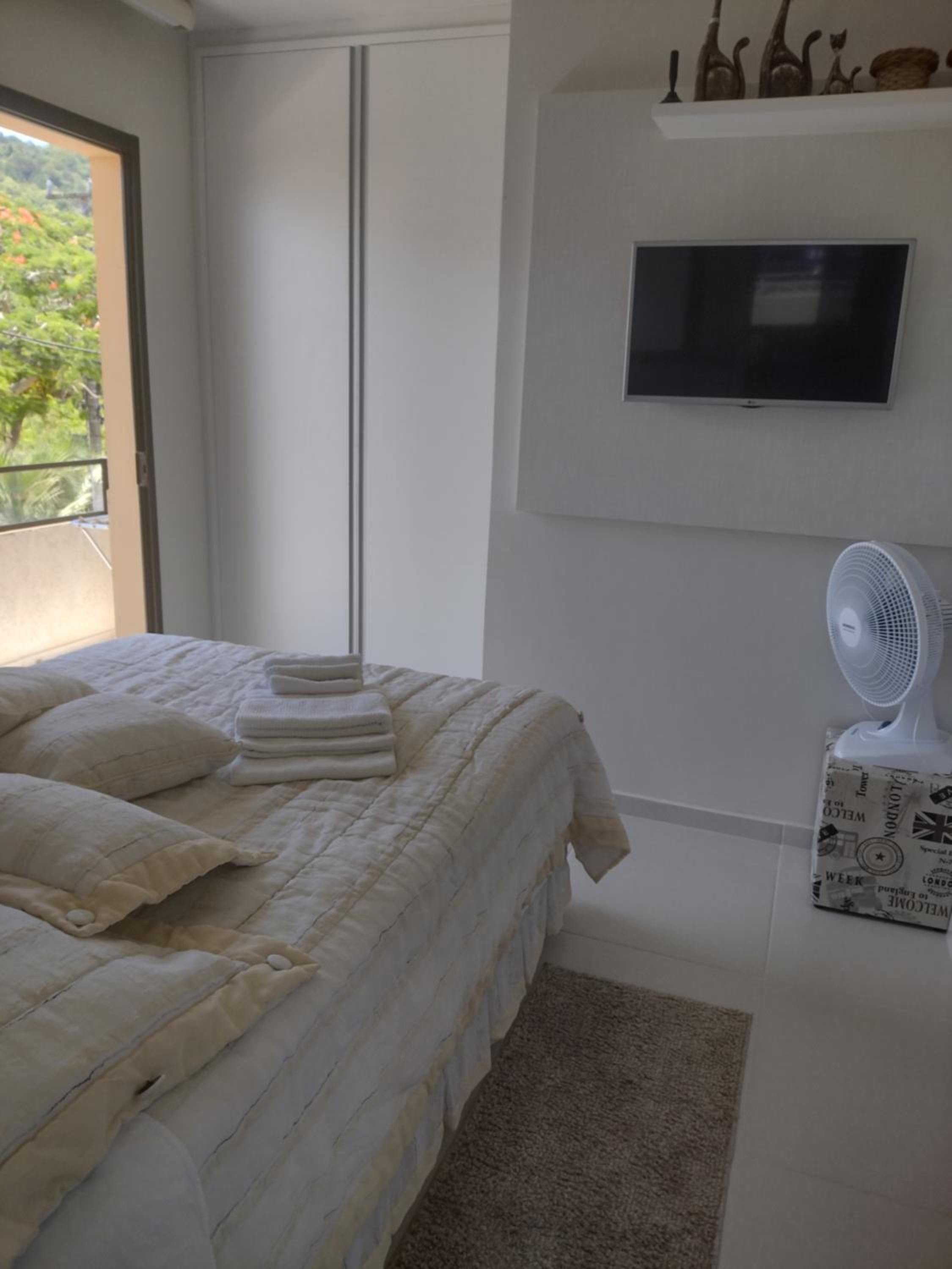 Quarto do Apto 4: cama de casal com toalhas dobradas, ventilador e TV na parede